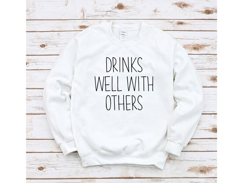 Może przedstawiać: Biała bluza z czarnym napisem "Drinks well with others".