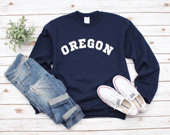 Può includere: Una felpa blu navy con la scritta "OREGON" stampata in bianco sul davanti. La felpa è stesa su una superficie di legno bianco con un paio di scarpe da ginnastica bianche e un paio di jeans blu.