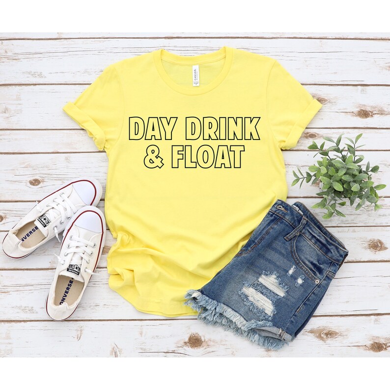 Op de afbeelding: Geel T-shirt met de tekst "DAY DRINK & FLOAT" in zwarte omrande letters. Het shirt ligt op een wit houten oppervlak met een paar witte sneakers en een spijkerbroek.