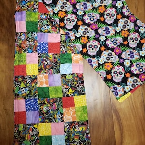 May include: A quilted table runner featuring a patchwork design with various colorful squares. One side showcases a black fabric with sugar skulls and flowers. The other side has a patchwork design with paisley and skull patterns.