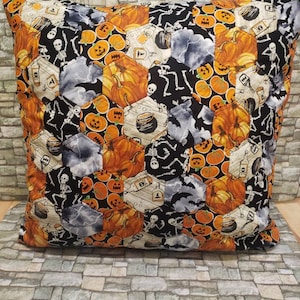 May include: Square Halloween cushion with a patchwork design. The fabric features orange pumpkins, skeletons, bats, and potion bottles on a black background. The cushion is placed on a stone-like surface.