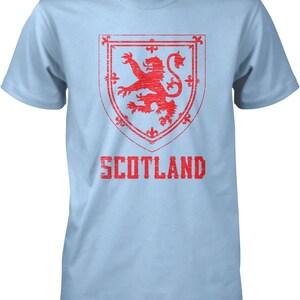 May include: A light blue t-shirt with a red distressed graphic of the Scottish coat of arms, a lion rampant within a shield, and the word "SCOTLAND" printed below.