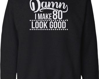 May include: A black long-sleeve crewneck sweatshirt featuring white text that reads "Damn I MAKE 80 LOOK GOOD" with a decorative line of dots separating the phrases. The design is centered on the chest of the garment.