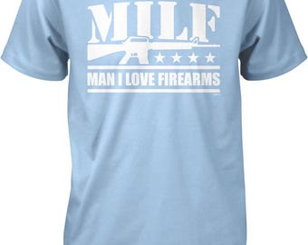 May include: A light blue short-sleeved t-shirt featuring a white graphic print on the chest. The design includes the text MILF, a silhouette of a rifle, four stars, and the text MAN I LOVE FIREARMS centered below the rifle graphic.