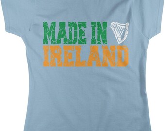May include: A light blue short-sleeve t-shirt featuring the text "MADE IN" in distressed green letters, followed by a white harp icon, and the word "IRELAND" in distressed orange letters centered on the chest.