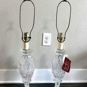 May include: A pair of clear crystal table lamps with ornate designs. The lamps have a square base and a tall, slender body with a decorative pattern. The lamps are topped with a simple, curved metal shade holder.