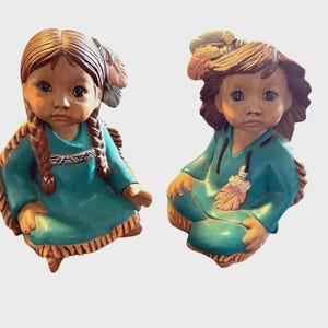 May include: Two ceramic figurines of Native American children. Each child is wearing a teal-colored tunic and has brown hair. One child has braids, and the other has loose curls. Both have feather adornments.