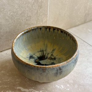 May include: A ceramic bowl with a rounded shape. The bowl has a mix of blue, yellow, and brown colours, with a dark centre. The rim has a darker brown hue. This bowl is suitable for serving food, and is handmade.