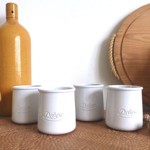Puede incluir: Cuatro recipientes de cerámica blancos con la palabra "Deluxe" en relieve, dispuestos sobre una superficie tejida. Una botella marrón grande y una tabla de cortar de madera en el fondo. Diseño sencillo y clásico.