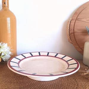 Può includere: Un piatto in ceramica color crema con un bordo largo con un motivo di rettangoli bordeaux e neri alternati. Il piatto è appoggiato su una superficie tessuta, con una bottiglia marrone e fiori sullo sfondo.