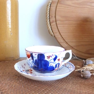 Op de afbeelding: Een witte kop en schotel met een bloemenpatroon in blauw, rood en paars. De kop heeft een gebogen oor en staat op een bijpassende schotel. De set staat op een geweven mat.