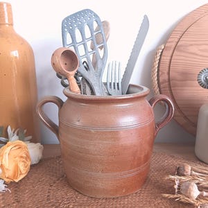 May include: A rustic, brown ceramic jar with two handles, filled with kitchen utensils. The jar is surrounded by other kitchen items, including a wooden cutting board, a bottle, and a small white jar with kitchen tools.