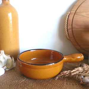 May include: A small, round, mustard-yellow ceramic bowl with a handle and a black rim. The bowl is empty and sits on a textured surface, with a garlic bulb and other kitchen items in the background.