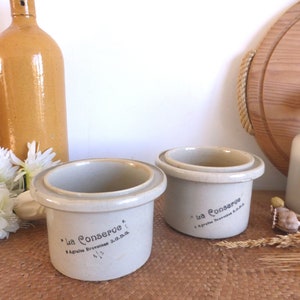 May include: Two vintage ceramic jars with lids, both labeled "La Conserve" with the text "Agriculteurs Brevetée S.D.G. 1/2 L." in black lettering. The jars are a light gray color and have a rustic, farmhouse aesthetic.