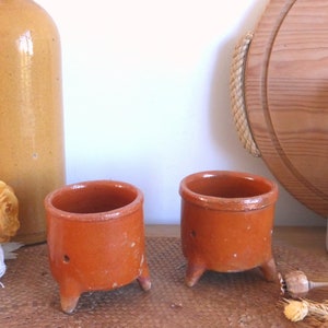 Könnte beinhalten: Zwei terracottafarbene Eierbecher mit jeweils drei Beinen. Die Becher stehen auf einer braunen Oberfläche.