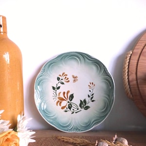 May include: A vintage ceramic plate with a scalloped edge and a floral design in brown, green, and white. The plate is displayed with a brown bottle and other decorative items, creating a rustic aesthetic.