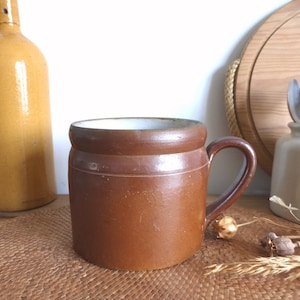 May include: A brown ceramic mug with a white interior and a handle. The mug has a rustic, vintage look.