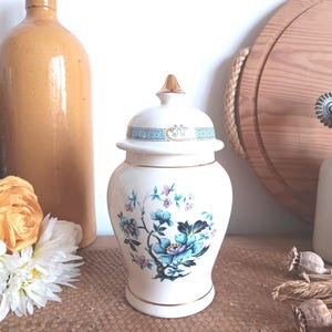 Könnte beinhalten: Weiße Keramikvase mit goldenem Deckel und Blumenmuster. Die Vase hat einen blauen und goldenen Rand um den Deckel und ein Blumenmuster mit blauen, rosa und grünen Blumen auf dem Korpus.