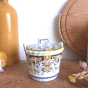 Może przedstawiać: Biały ceramiczny słoik z niebieskim i żółtym wzorem kwiatowym. Słoik ma pokrywkę z uchwytem i małym dzióbkiem. Słoik stoi na drewnianej powierzchni.