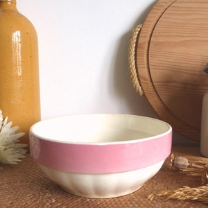 Puede incluir: Un tazón de cerámica blanco con una banda rosa alrededor del borde. El tazón está sobre una superficie tejida.