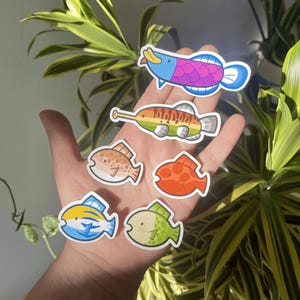 Puede incluir: Seis pegatinas de peces de colores con diferentes diseños. Las pegatinas están en una mano, y la mano está sosteniendo las pegatinas contra una planta verde.