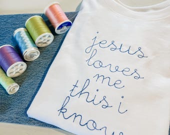Puede incluir: Camiseta blanca con bordado azul que dice "Jesús me ama, esto lo sé".