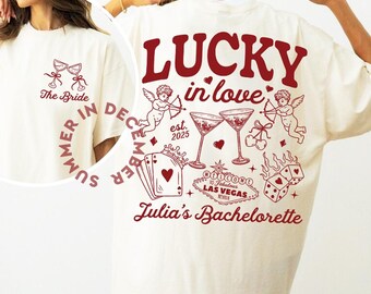 May include: Cream-colored t-shirt with a burgundy graphic. The front features two martini glasses and the text "The Bride". The back displays "LUCKY in love", cherubs, martini glasses, playing cards, dice, and "Julia's Bachelorette" with "Las Vegas" text.