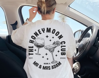 May include: White t-shirt with a black circle graphic that says "The Honeymoon Club" with a wine glass and a margarita glass inside the circle. The text below the circle says "Est. 2024 Miami, FL Mr. & Mrs. Adam"
