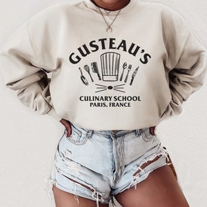 May include: A beige sweatshirt with a black graphic print of a chef's hat and kitchen utensils. The text "Gusteau's Culinary School Paris, France" is printed below the graphic.