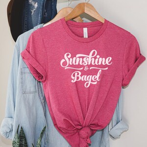 K&ouml;nnte beinhalten: Ein pinkfarbenes T-Shirt mit dem Text "Sunshine & Bagel" in wei&szlig;en Buchstaben.
