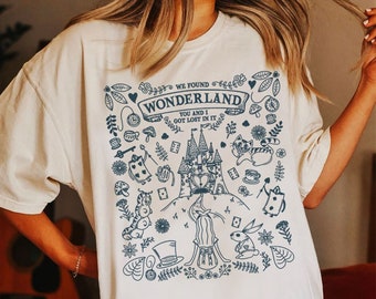 May include: A white t-shirt with a blue graphic print of a castle, a caterpillar, a rabbit, a teacup, a playing card, and other whimsical elements. The text "We found Wonderland, You and I got lost in it" is printed on the shirt.