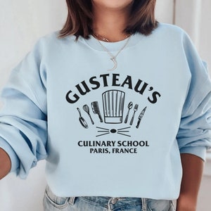 May include: Light blue sweatshirt with a black graphic of a chef's hat and kitchen utensils. The text "Gusteau's Culinary School Paris, France" is printed below the graphic.