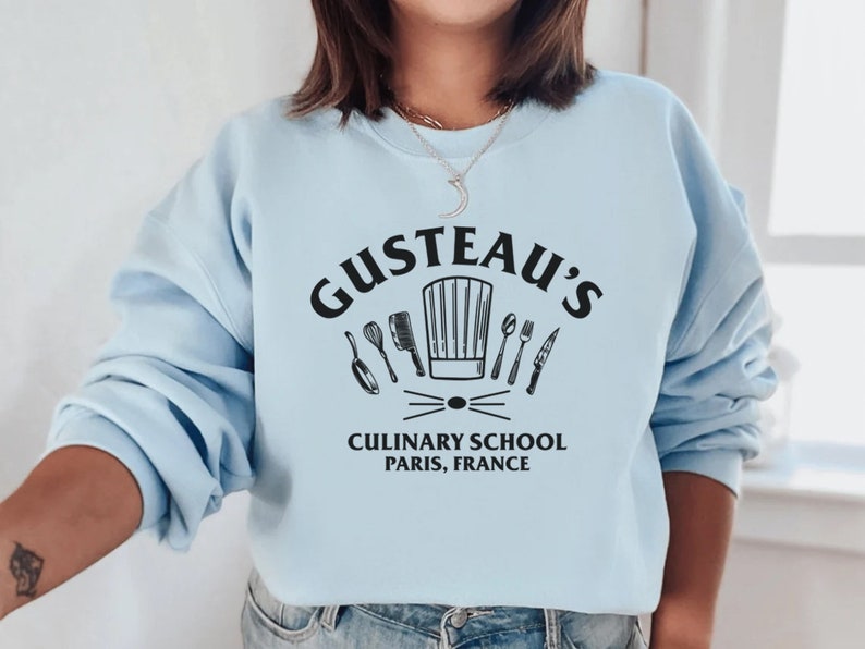 May include: Light blue sweatshirt with a black graphic of a chef's hat and kitchen utensils. The text "Gusteau's Culinary School Paris, France" is printed below the graphic.