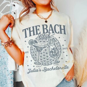 May include: Cream-colored t-shirt with the text "THE BACH Club" and "Julia's Bachelorette" in gray. The design includes illustrations of two cocktails and the year 2025. The shirt is a casual style.