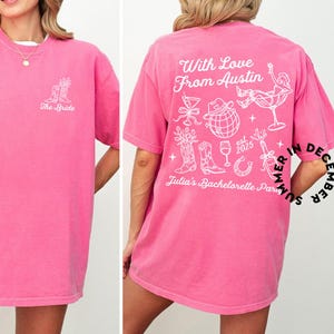 May include: Pink t-shirt with a white graphic design on the back. The design includes the text "With Love From Austin", a cowboy hat, a glass of wine, a horseshoe, and the text "Julia's Bachelorette Party". The front of the shirt has the text "The Bride" in white.