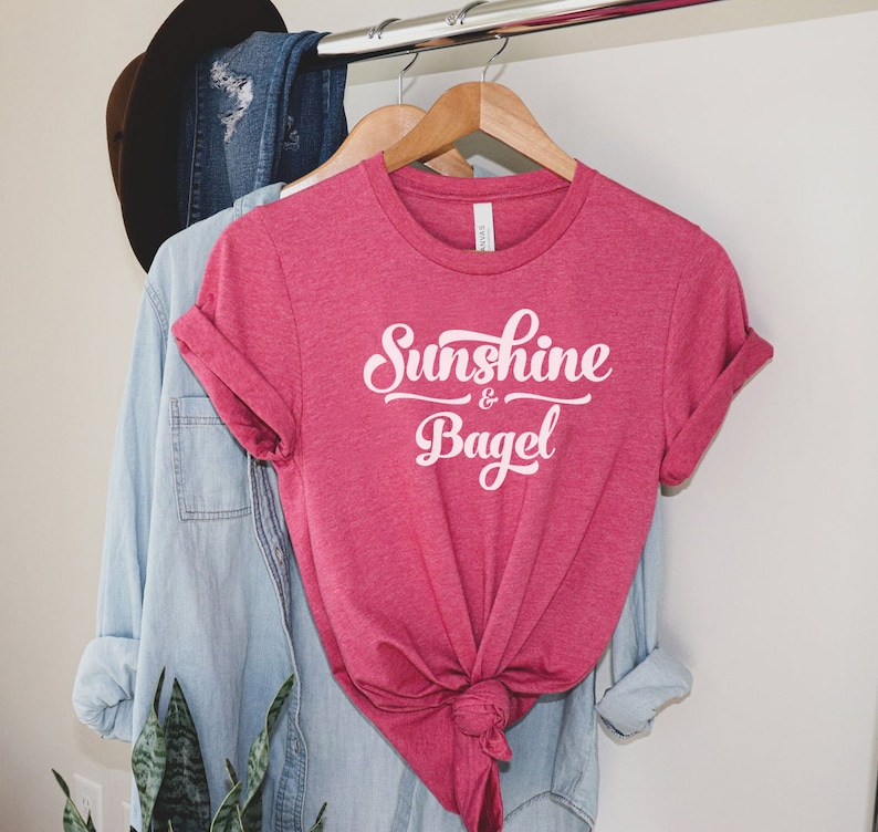 K&ouml;nnte beinhalten: Ein pinkfarbenes T-Shirt mit dem Text "Sunshine & Bagel" in wei&szlig;en Buchstaben.