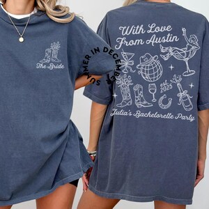 May include: A dark blue t-shirt with a white graphic design on the back. The design includes the text "With Love From Austin", a horseshoe, a bottle of champagne, a glass of wine, a cowboy hat, a disco ball, and a pair of cowboy boots. The text "Summer in December" is written in a circle around the design. The text "est. 2025" and "Julia's Bachelorette Party" are also included in the design. The front of the shirt has the text "The Bride" and a pair of cowboy boots.