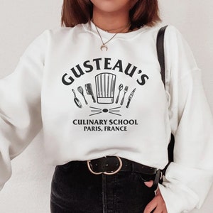 May include: White sweatshirt with black text that reads "Gusteau's Culinary School Paris, France". The text is surrounded by a chef's hat and kitchen utensils.