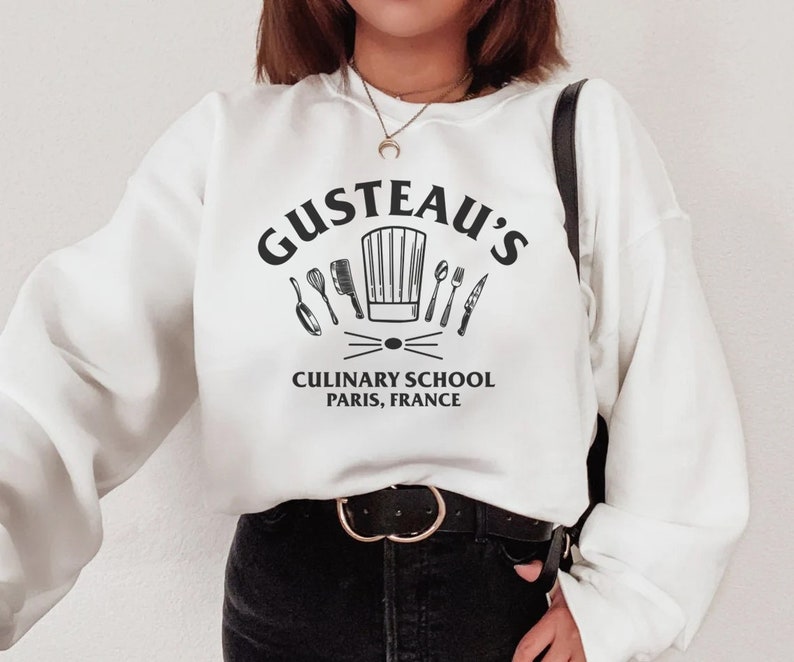 May include: White sweatshirt with black text that reads "Gusteau's Culinary School Paris, France". The text is surrounded by a chef's hat and kitchen utensils.