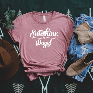 K&ouml;nnte beinhalten: Ein staubiges rosafarbenes T-Shirt mit dem Text "Sunshine & Bagel" in wei&szlig;en Buchstaben. Das Shirt liegt auf einer gr&uuml;n-wei&szlig;en gemusterten Oberfl&auml;che mit einem braunen Hut, einer blauen Jeans und braunen Stiefeln.