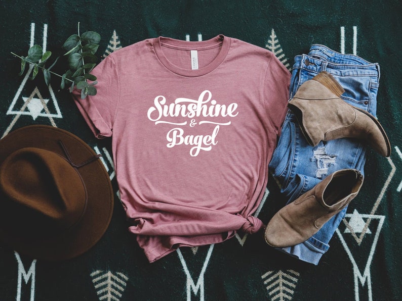 K&ouml;nnte beinhalten: Ein staubiges rosafarbenes T-Shirt mit dem Text "Sunshine & Bagel" in wei&szlig;en Buchstaben. Das Shirt liegt auf einer gr&uuml;n-wei&szlig;en gemusterten Oberfl&auml;che mit einem braunen Hut, einer blauen Jeans und braunen Stiefeln.