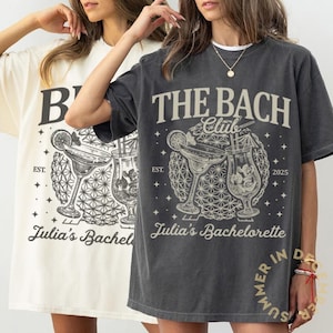 May include: Two oversized t-shirts, one cream and one charcoal, featuring a cocktail graphic. The charcoal shirt reads "THE BACH Club" with "Julia's Bachelorette" below. Both shirts have a vintage aesthetic, perfect for a bachelorette party.
