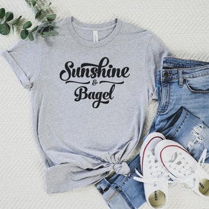 K&ouml;nnte beinhalten: Ein graues T-Shirt mit dem Text "Sunshine & Bagel" in schwarzer Schrift. Das Shirt liegt auf einer wei&szlig;en Oberfl&auml;che mit einer blauen Jeans und wei&szlig;en Turnschuhen.