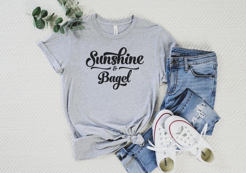 K&ouml;nnte beinhalten: Ein graues T-Shirt mit dem Text "Sunshine & Bagel" in schwarzer Schrift. Das Shirt liegt auf einer wei&szlig;en Oberfl&auml;che mit einer blauen Jeans und wei&szlig;en Turnschuhen.