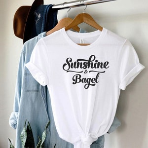 K&ouml;nnte beinhalten: Wei&szlig;es T-Shirt mit schwarzem Text in kursivschrift, der "Sunshine & Bagel" lautet.