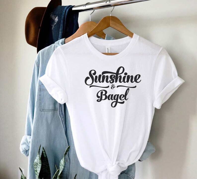 K&ouml;nnte beinhalten: Wei&szlig;es T-Shirt mit schwarzem Text in kursivschrift, der "Sunshine & Bagel" lautet.
