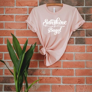 K&ouml;nnte beinhalten: Ein hellrosa T-Shirt mit dem Text "Sunshine & Bagel" in wei&szlig;en Buchstaben vor einem Hintergrund aus Ziegeln.