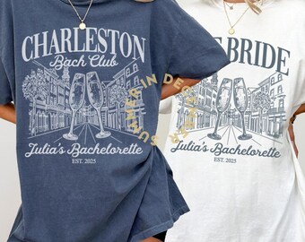 May include: Two oversized t-shirts with a city skyline graphic and the text "Charleston Bach Club" and "Bride" with the words "Julia's Bachelorette" and "EST. 2025" below the graphic.