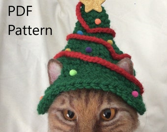 May include: A brown and orange cat wearing a green knitted Christmas tree hat with red yarn and colorful pom-poms. The hat has a gold star on top. The cat is looking at the camera. PDF Pattern.