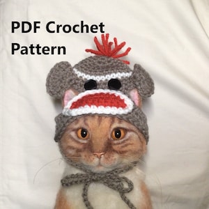 May include: A cat wearing a handmade crochet sock monkey hat. The hat is gray, white, and red, with black button eyes and orange yarn accents. The text "PDF Crochet Pattern" is in the upper left corner.